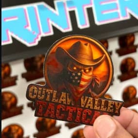 A hand holding a freshly printed Outlaw Valley Tactical sticker over a full sheet of them.