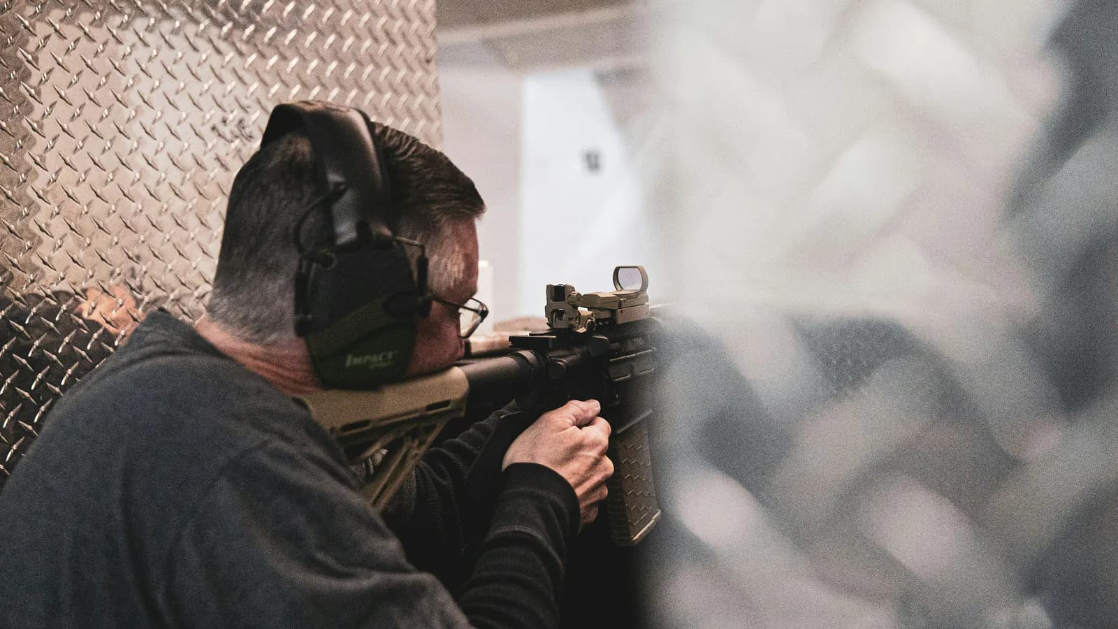 A shooter in electronic ear muffs shoulders an AR-15 fitted with a tan reflex red dot sight and a folded backup iron sight inside an indoor range lane