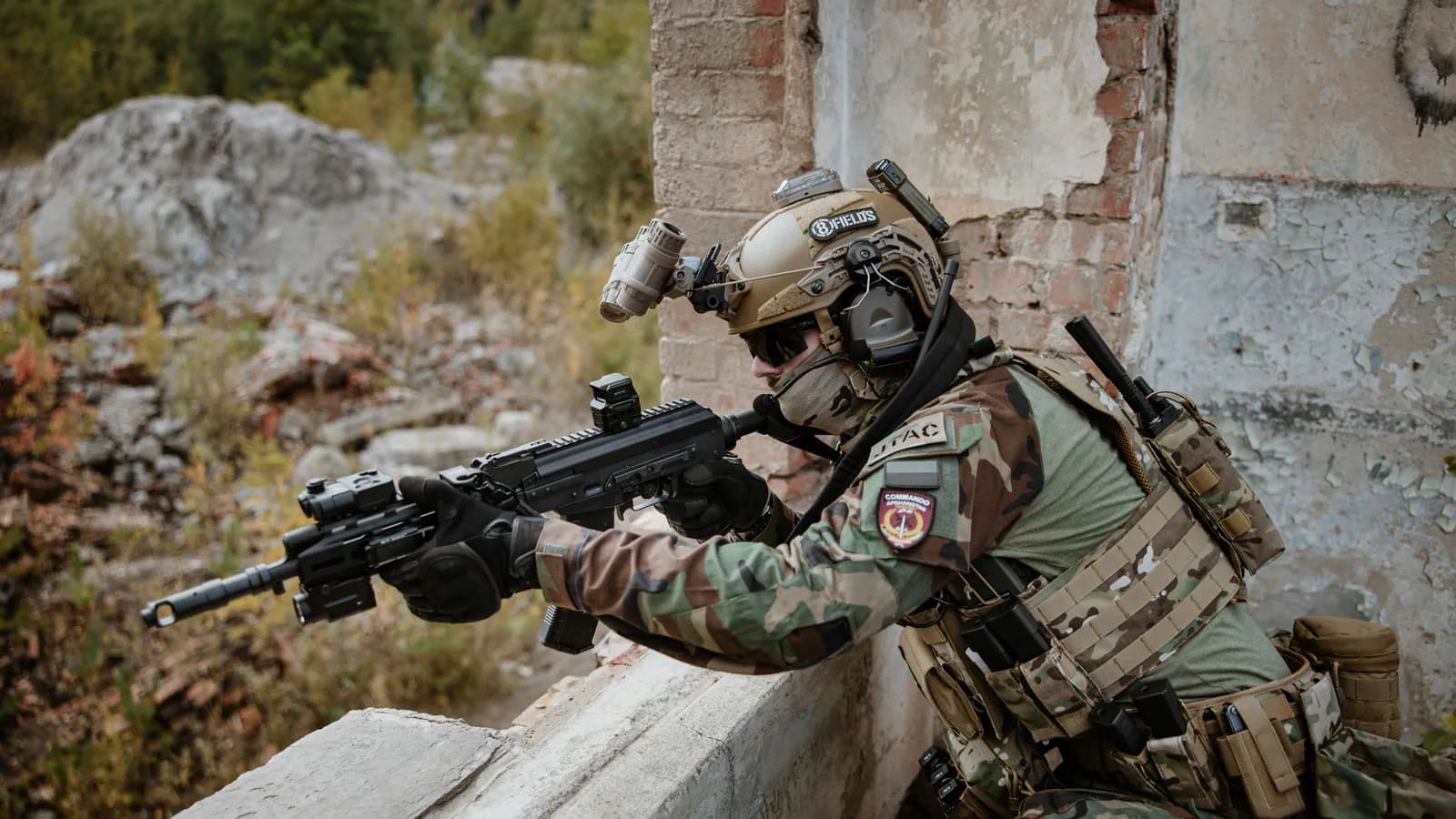 A shooter braces a carbine over a low wall, the rifle carrying a small enclosed red dot on the receiver and a second magnifying optic further forward on the rail