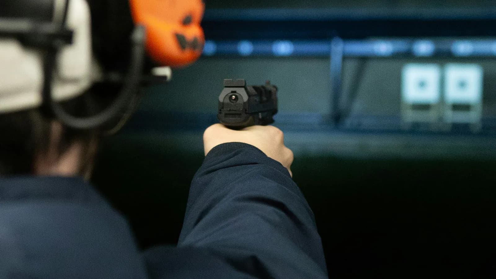 A shooter in a white cap and orange ear protection extends a pistol in a two handed grip toward brightly lit paper targets on a dark outdoor range at night