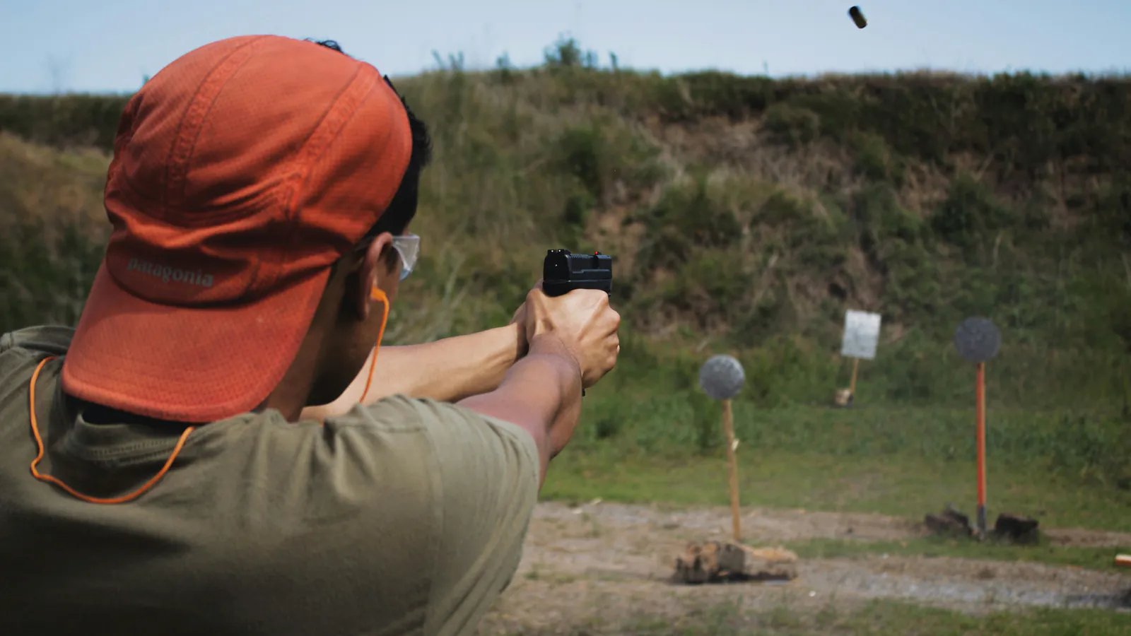 A shooter in an orange cap fires a pistol with a two handed grip at an outdoor range as a spent case flies clear of the slide, with steel and paper targets set along the berm