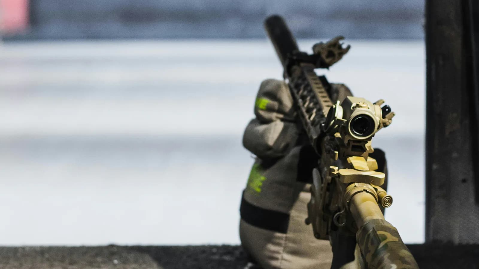 An AR-15 in multicam and flat dark earth rests on a support bag at a shooting bench, fitted with a magnified optic and a small red dot mounted at a 45 degree offset