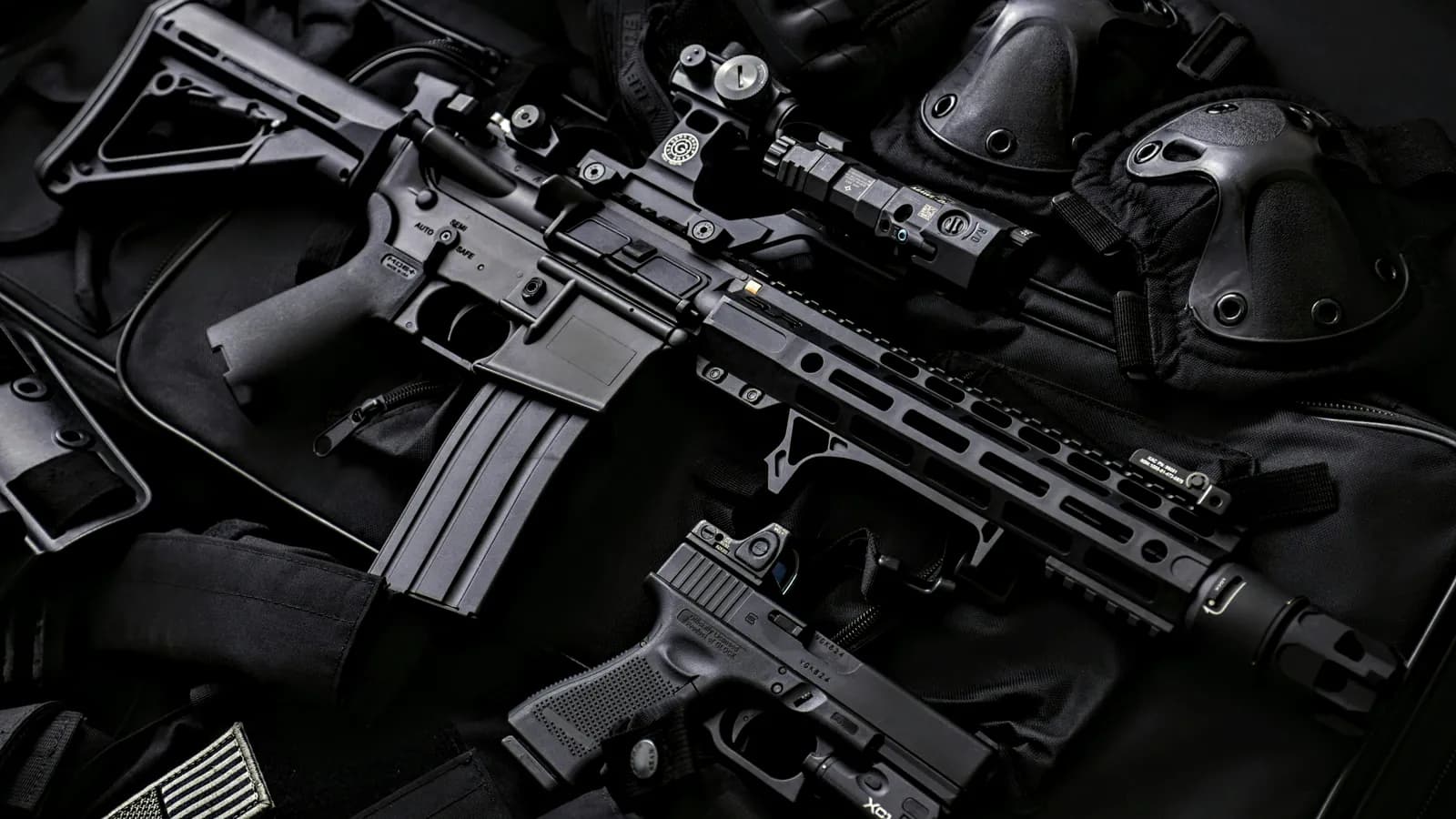 An AR-15 laid out on dark gear beside a pistol, a weapon light, a red dot sight and its mount, with the receiver, magazine and slotted handguard all in view