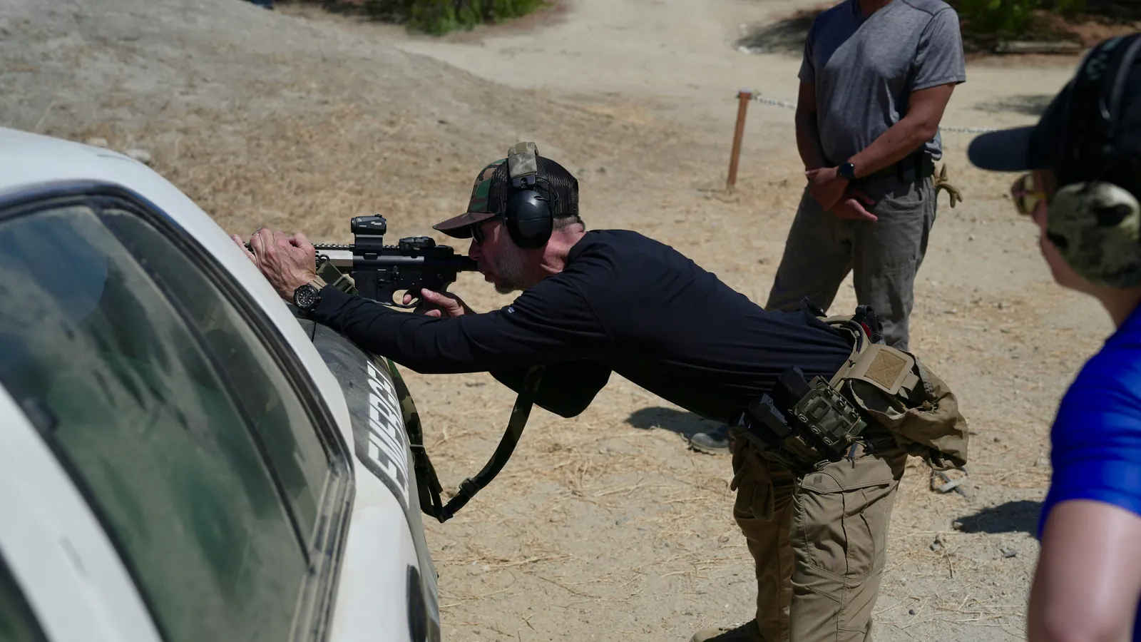 A shooter in a camo cap and electronic ear muffs braces a rifle over the hood of a vehicle on a dusty outdoor range while an instructor stands by on the firing line