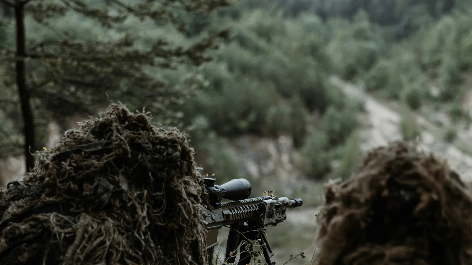Two shooters in ghillie suits lie behind a bipod mounted rifle with a large magnified scope, looking down a wooded valley at a track running into the distance