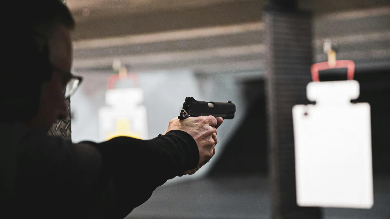 A shooter in ear protection and safety glasses fires a semiautomatic pistol with both hands at a paper target in an indoor range lane
