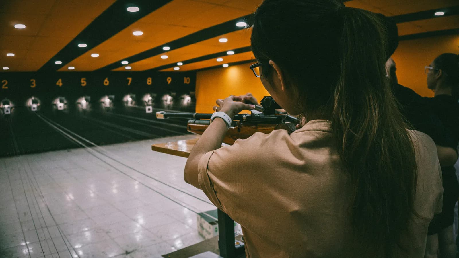 A shooter aims a rifle down a numbered indoor range lane toward a row of paper bullseye targets