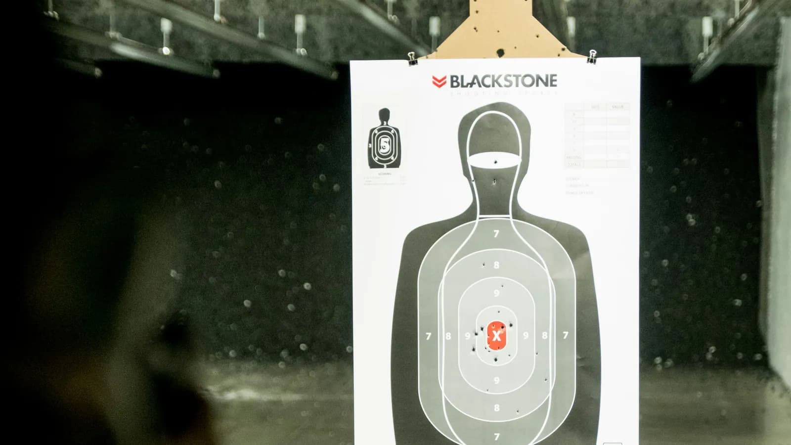 A paper silhouette target hangs in a dim indoor range lane with its scoring rings and red center mass marker clearly visible, a shooter blurred in the foreground