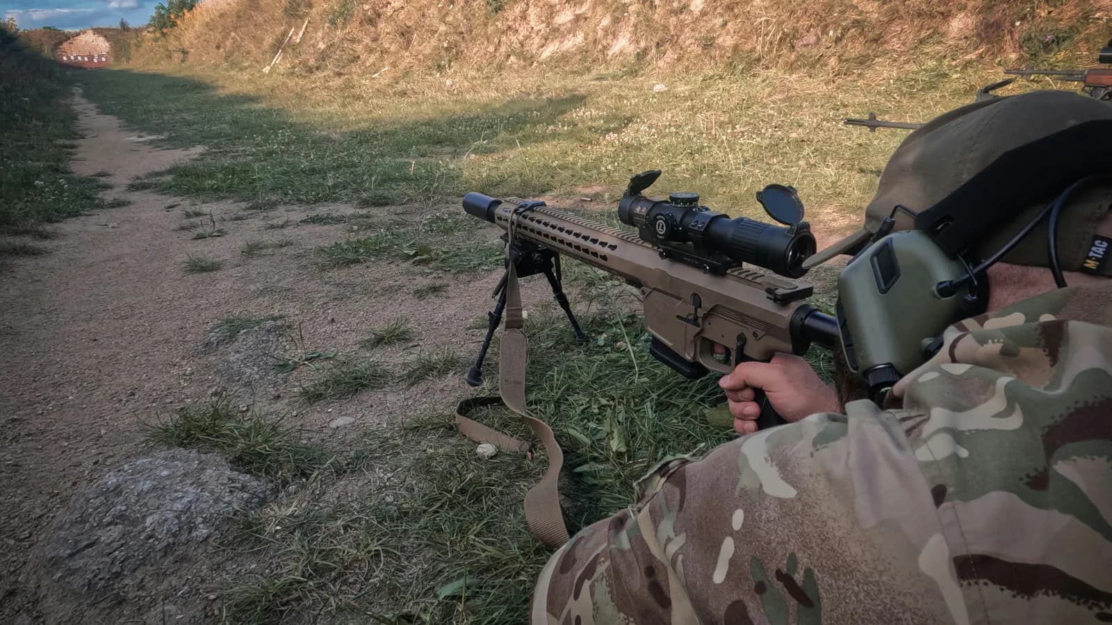 A tan precision rifle rests on a bipod with a large magnified scope mounted above the receiver as a shooter in multicam settles in behind it at an outdoor range