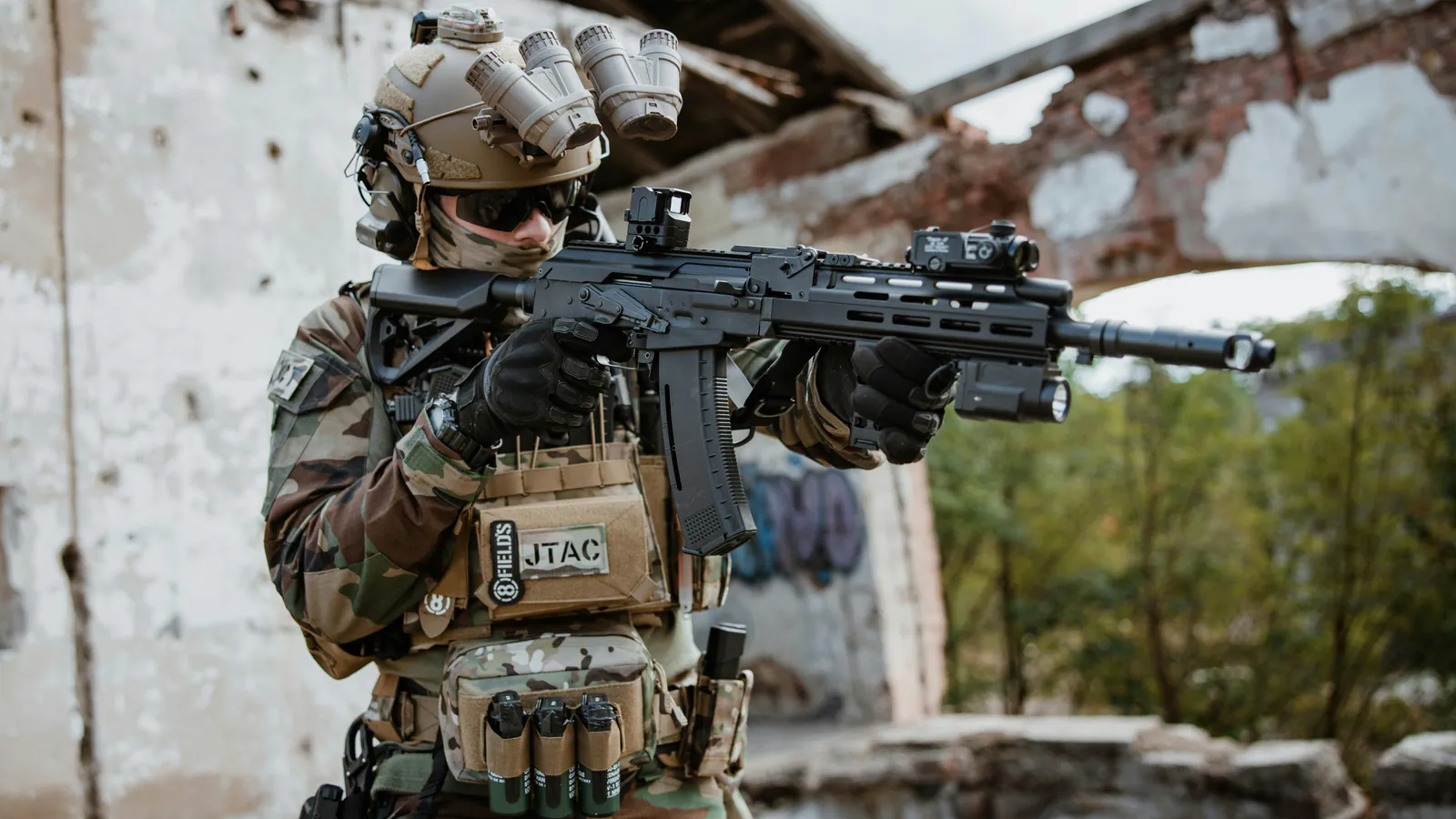A shooter in a helmet with four tube night vision goggles flipped up above his eyes shoulders a carbine fitted with a red dot sight and a weapon light