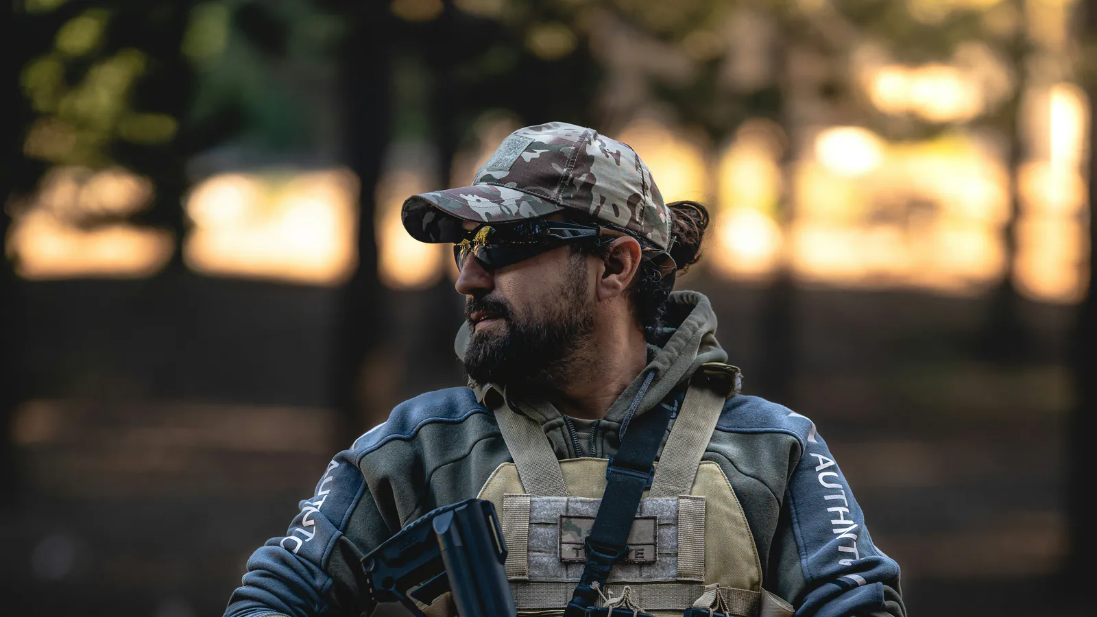 A bearded shooter in a camo cap, wraparound eye protection and a plate carrier stands on an outdoor range with a rifle across his chest as low sun burns through the trees behind him