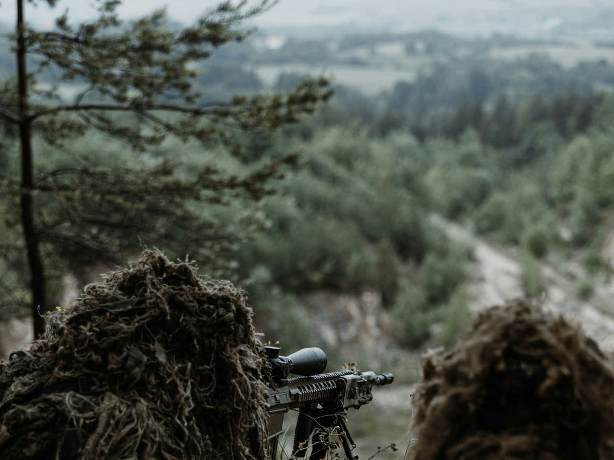 Two shooters in ghillie suits behind a scoped rifle on a ridge